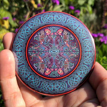 A circular Celtic design featuring four men and four birds facing left and right, then the same but upside down, sits in the centre of the design. Colours are blues, reds, pinks and purples. A wide Celtic knot border in shades of blue with a red edge surrounds the central design.
