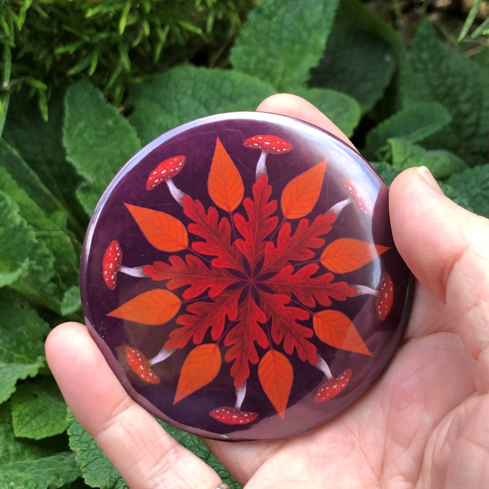 A mandala-style painting featuring a circle of red oak leaves in the centre, followed by a circle of simple orange leaves outside of that, and finally a circle of red and white-dotted toadstools on the outer edge. The background is purple.