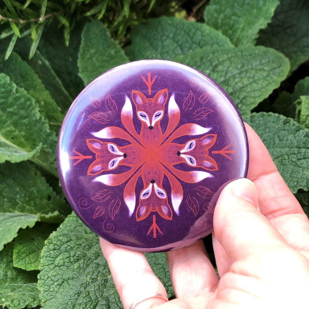 A mandala-style painting featuring a circle of four red-brown fox heads with white faces. The foxes face towards the centre. White-tipped tails curl out in the gaps between the foxes. The foxes are surrounded by a less prominent outer mandala of red-brown leaves and twigs. The background is purple.