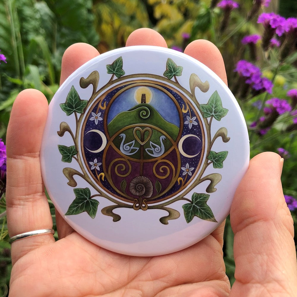 A circular design on a white background featuring the Chalice Well (Vesica Pisces) symbol inside a circle of ivy leaves. Glastonbury Tor, the sun rising behind the tower against a blue sky, nestles inside the upper circle of the symbol, with two swans in the area where the two circles join. An ammonite is inside the lower circle, against a maroon background. To the left and right are two opposing crescent moons, and small white flowers.