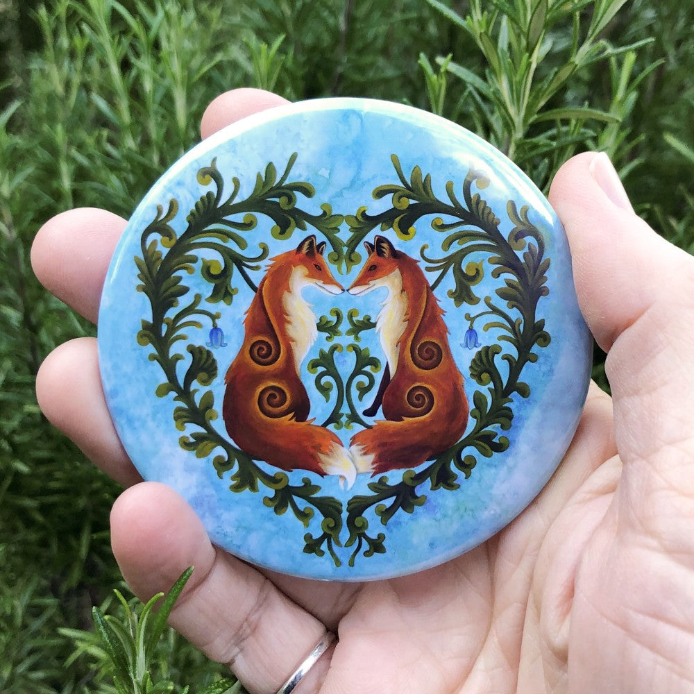 A symmetrical design. Two red foxes sit facing each other, noses touching and tails entwined. Surrounding the foxes are two green, leaved branches which form the two halves of a heart shape. A single bluebell flower hangs behind each fox. The background is a mottled blue.