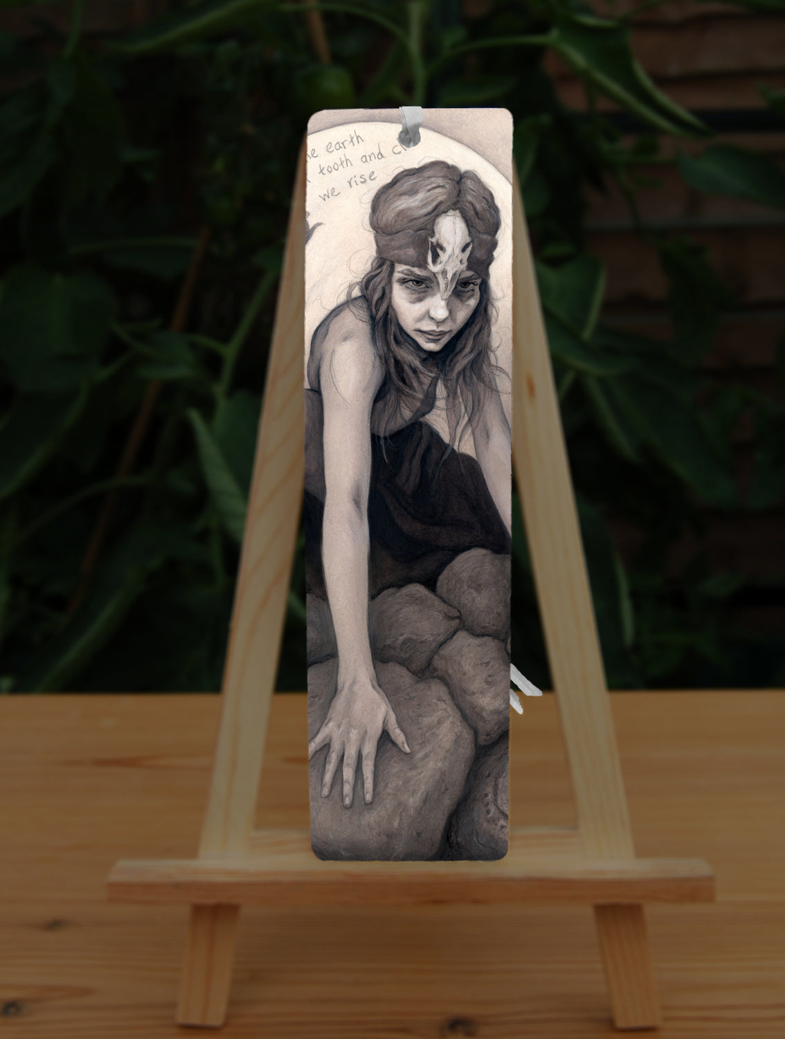 A tall bookmark: A young woman crouches on a pile of stones, her hands resting on stones in front of her. The woman wears a headband with a raven skull attached to the front. A full moon rises behind her.