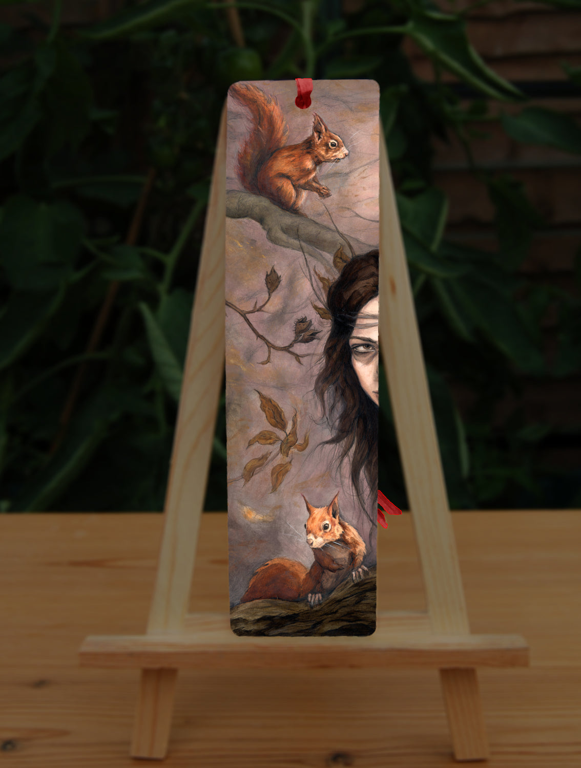 A tall bookmark: One side of the face of a young woman peers in from the right. A red squirrel sits on a branch above her, with another below her. Autumnal leaves and branches fill the scene. Colours are reds, oranges and browns.