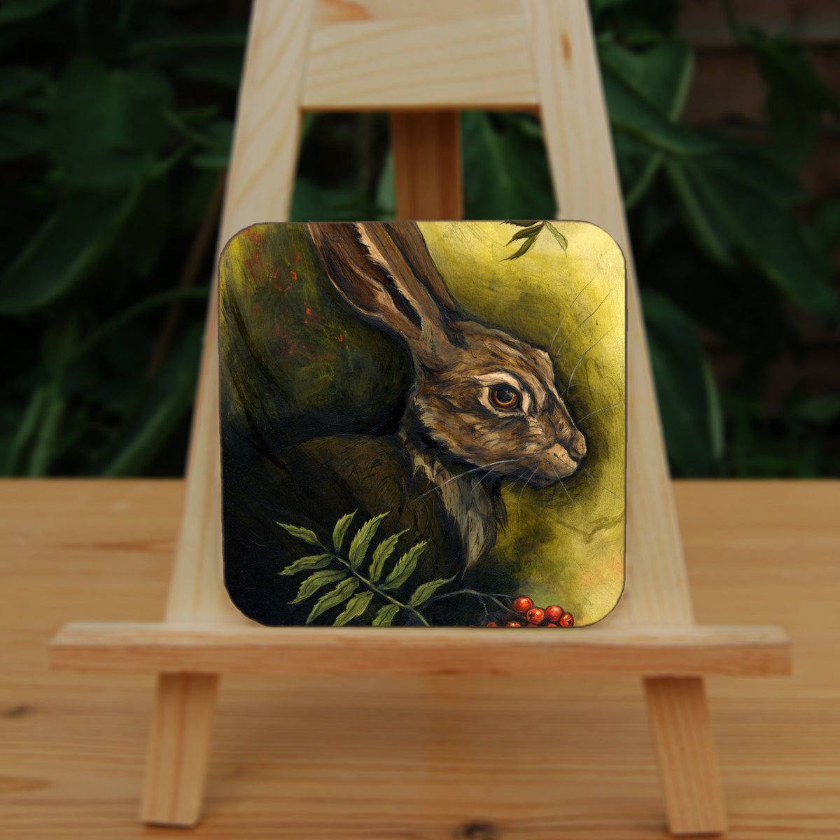 A square coaster: The head and shoulders of a hare facing to the right. Sprigs of red rowan berries and leaves are drawn below. The background is mottled yellows and browns.