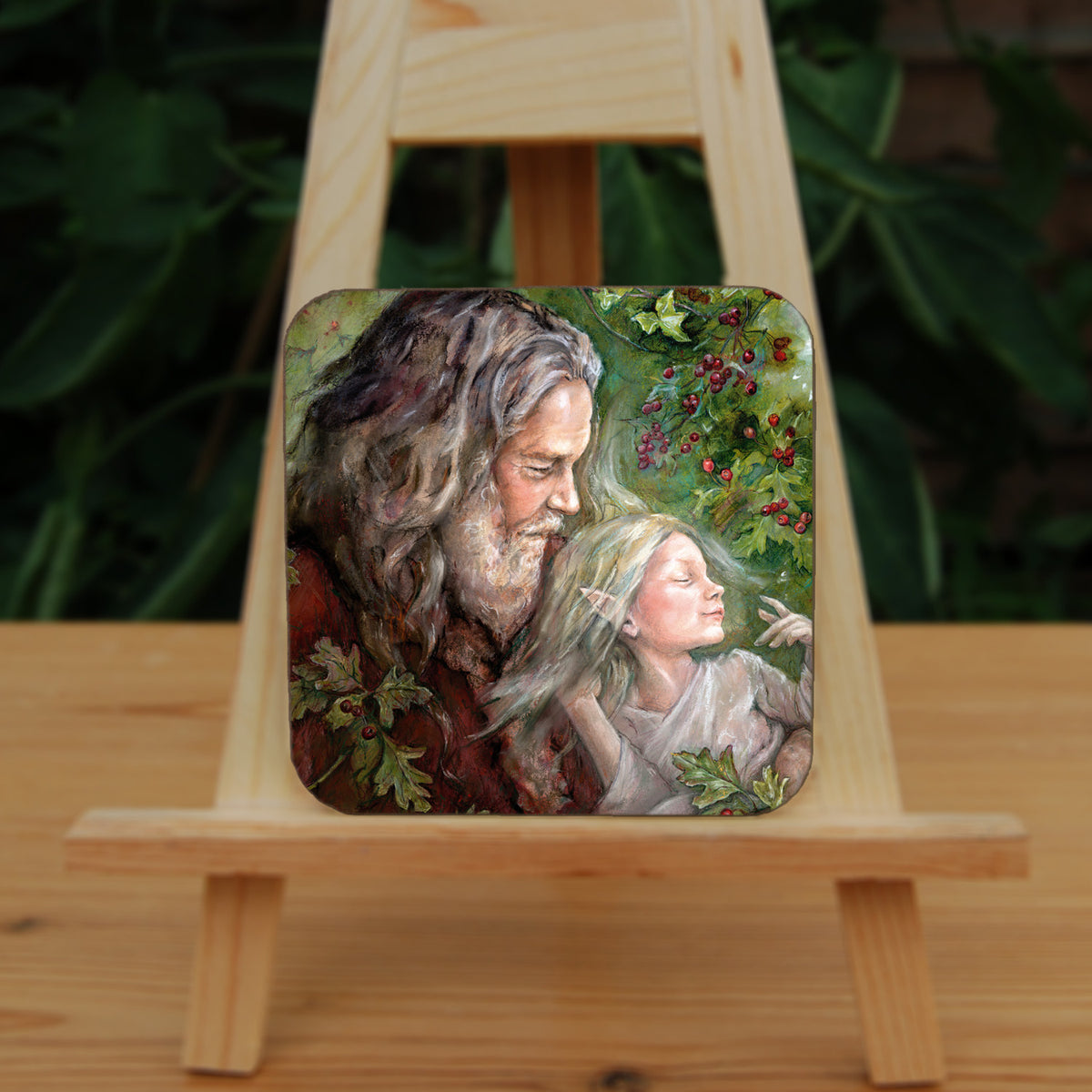 A square coaster: An older man with long hair, grey beard, and wearing a red coat cradles a sleeping fairy, wearing a pale gossamer dress, in his arms. Sprigs of berried hawthorn surround them. Branches of hawthorn and ivy fill the background.