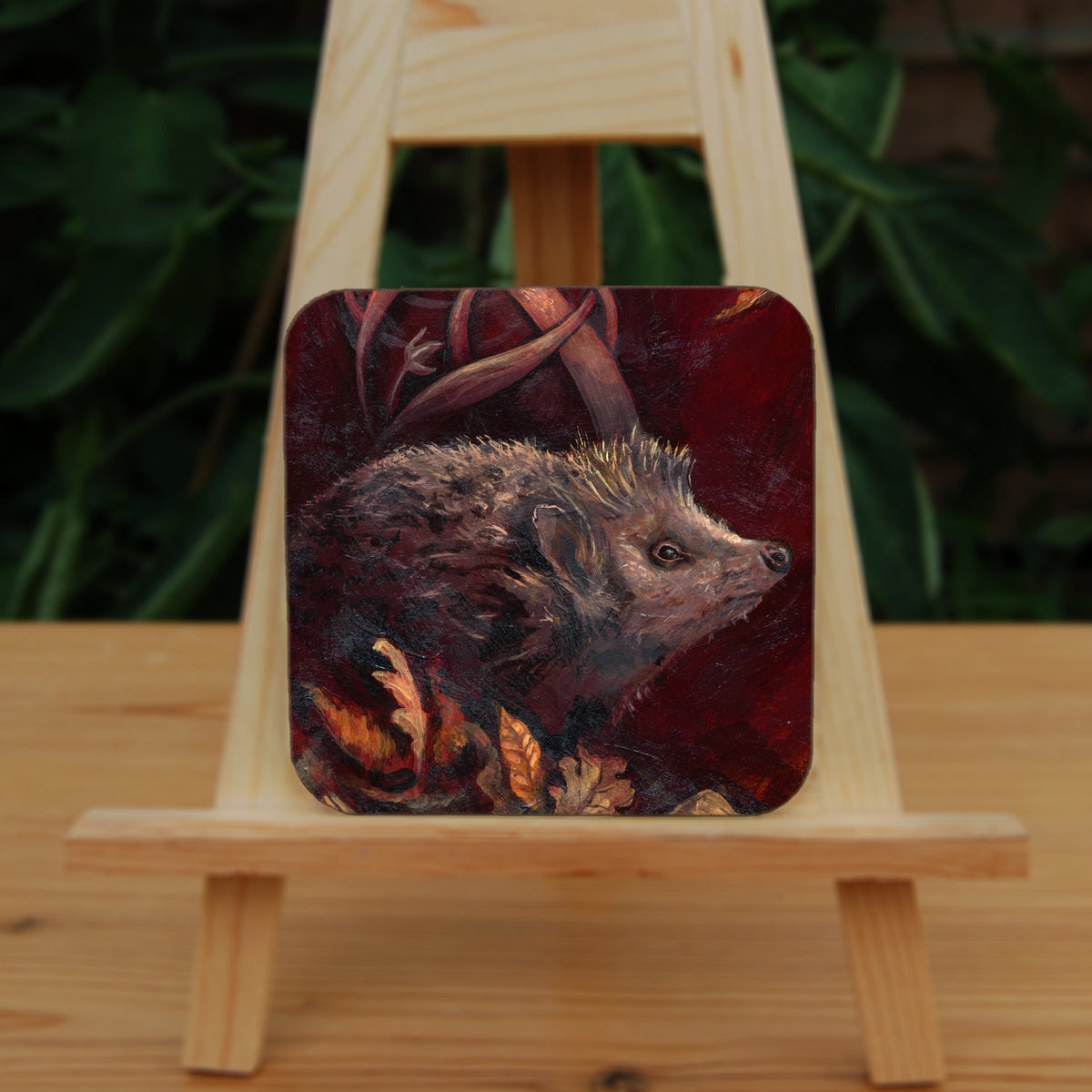 A square coaster: A hedgehog shuffles through crisp brown autumn leaves lying on the ground. Woven branches rise upwards, and a leaf falls towards the ground. Colours are browns and reds.
