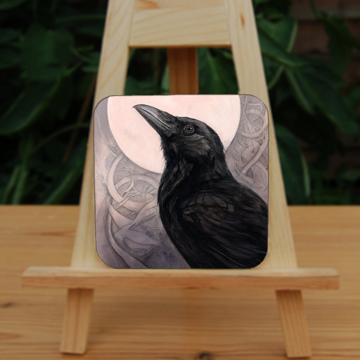Square coaster: The head and body of a raven standing facing towards the left, head tilted upward, behind which is a full moon. The background consists of swirls in a pale pink-brown colour.