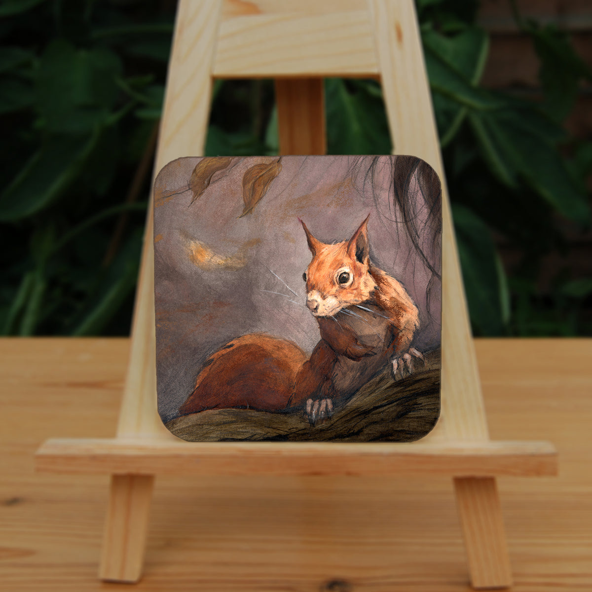 A square coaster: A red squirrel sitting on a branch facing the viewer. Autumnal leaves and branches fill the scene. Colours are reds, oranges and browns.