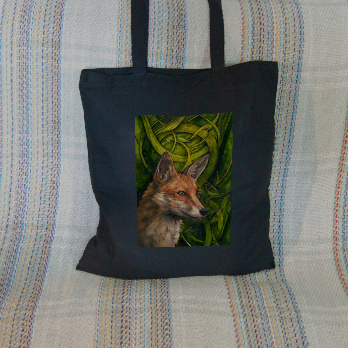 Black tote bag: The head and neck of a red fox, their face angled towards the front right. The background consists of swirls of branches and leaves in a rich green colour.