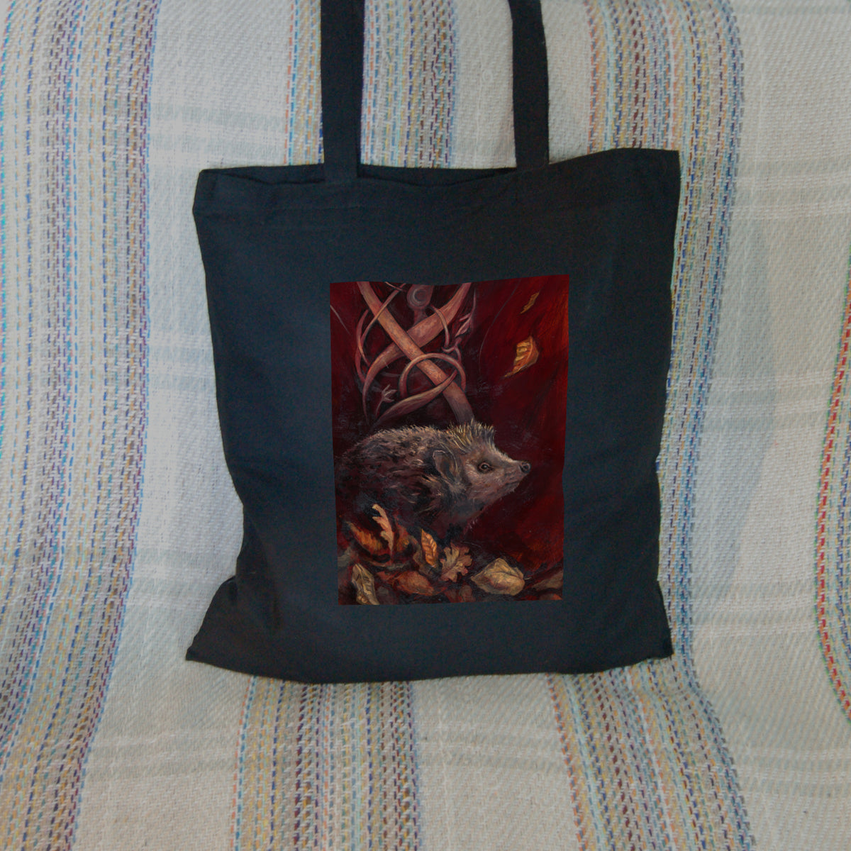 Black tote bag: A hedgehog shuffles through crisp brown autumn leaves lying on the ground. Woven branches rise upwards, and a leaf falls towards the ground. Colours are browns and reds.