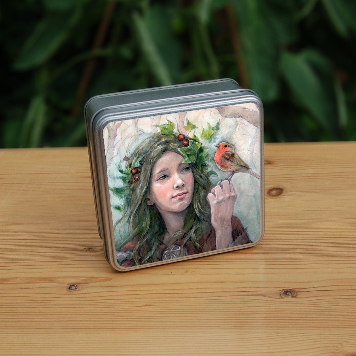 Square metal tin: A young woman looks towards a robin sitting at eye level on the fingers of her hand. She wears a red cloak with white fur trim, closed with a large Celtic brooch. Sprigs of red berries and ivy are woven into her hair. Bare branches of trees fill the background.