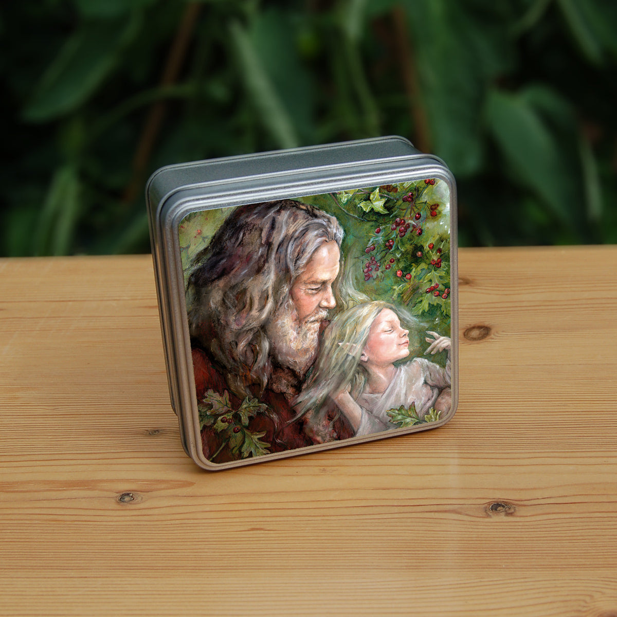 Square metal tin: An older man with long hair, grey beard, and wearing a red coat cradles a sleeping fairy, wearing a pale gossamer dress, in his arms. Sprigs of berried hawthorn surround them. Branches of hawthorn and ivy fill the background.
