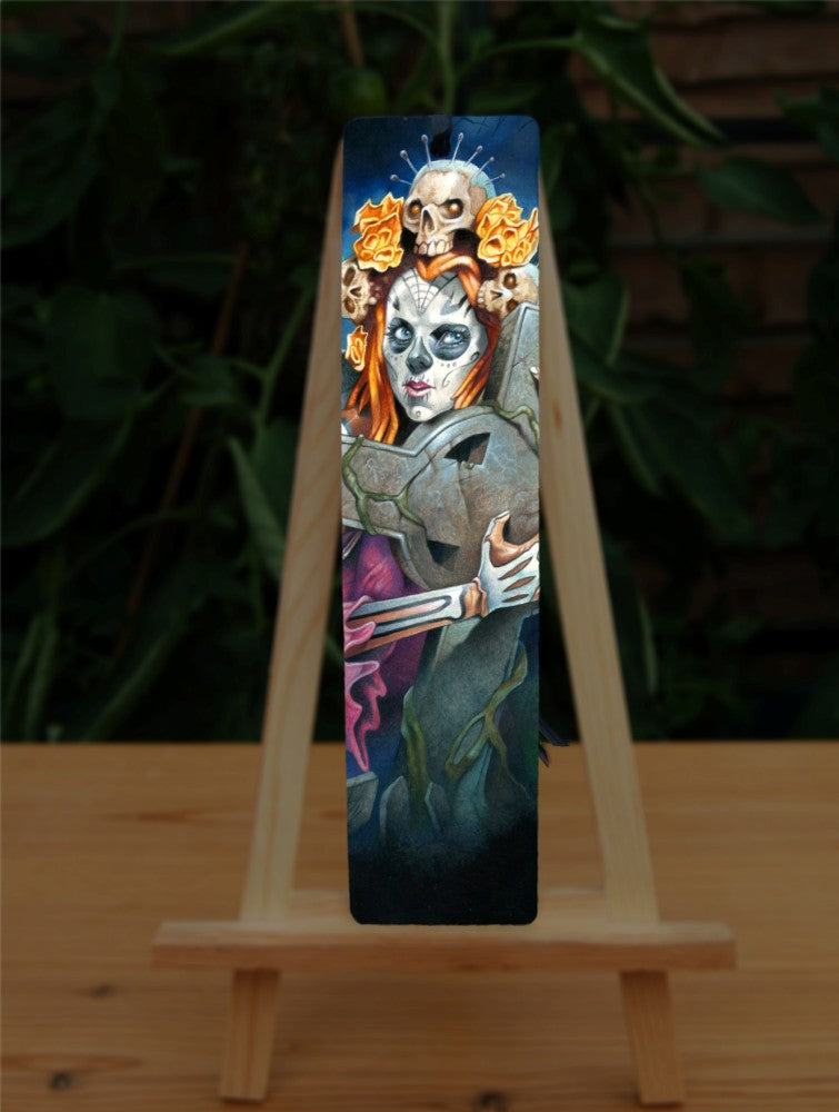 A woman with her face and arms decorated as a skeleton clutches a large stone Celtic cross to her chest. Her red hair is decorated with skulls with pins protruding from their heads.