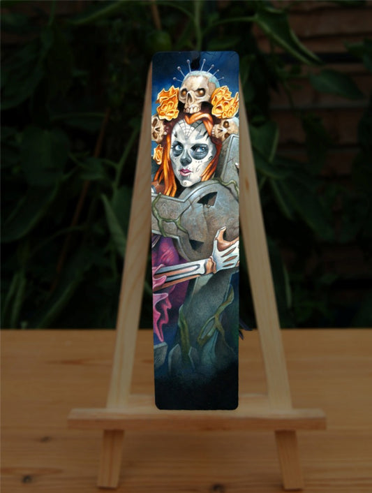 A woman with her face and arms decorated as a skeleton clutches a large stone Celtic cross to her chest. Her red hair is decorated with skulls with pins protruding from their heads.