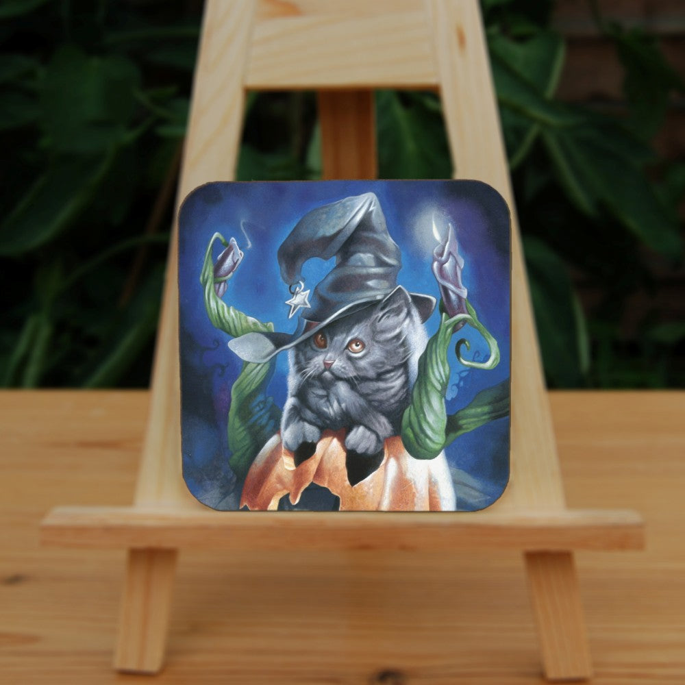 A grey cat or kitten sits on the top half of a broken Halloween pumpkin, his paws in the pumpkin's eyes. The cat is wearing a bent witch's hat with a star at the end. Green tendrils twist out of the pumpkin, one of them has a lit purple candle at the top.