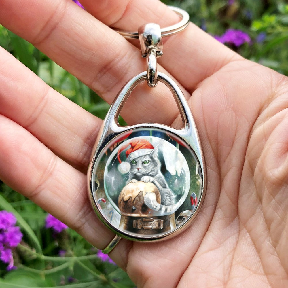 A pale silver tabby cat wearing a santa hat sits in a snow globe, front paws resting on a Christmas pudding. "Krampuss" is written on the globe stand beneath.