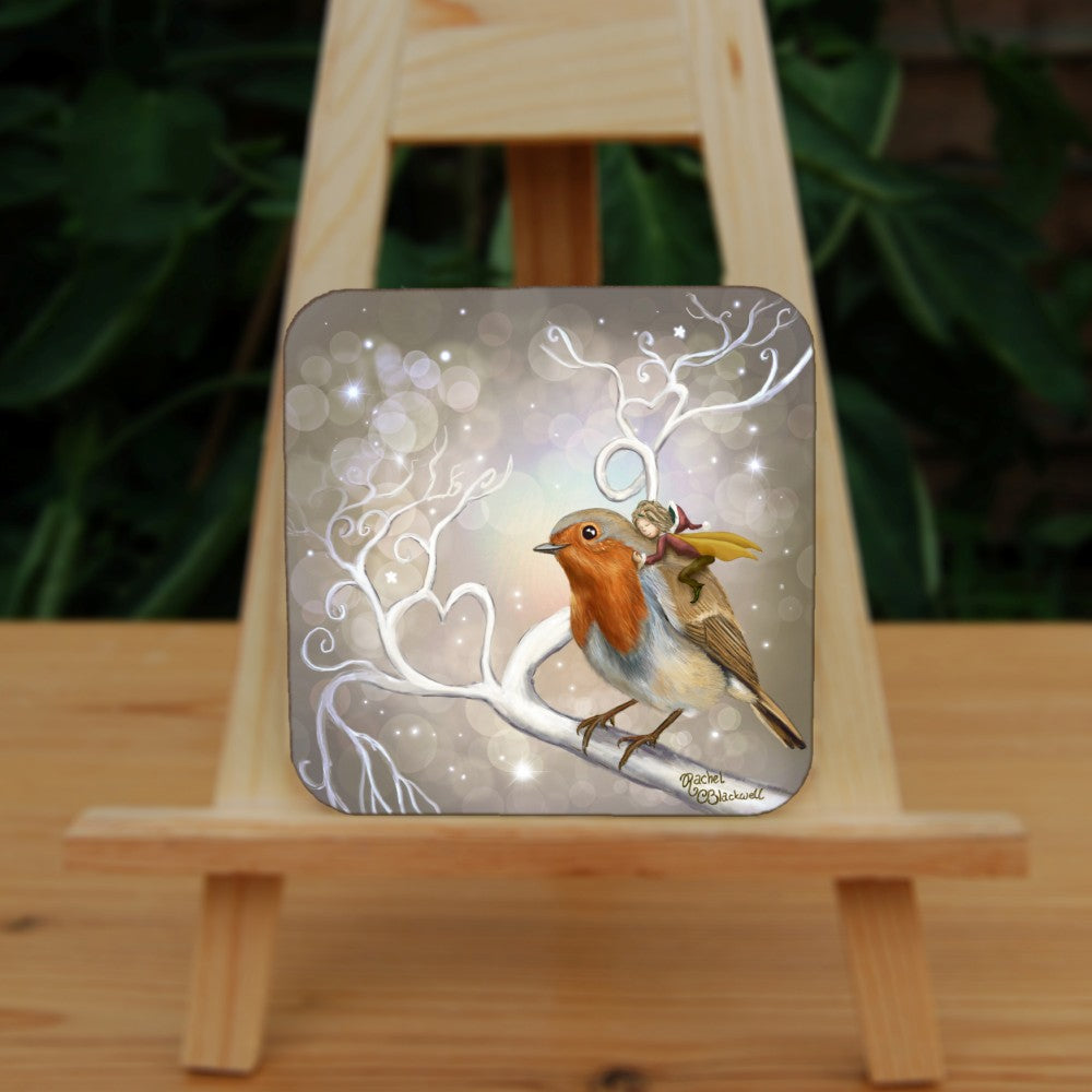 A robin perches on a white leafless branch. Twigs twist and curl from the branch, one forming a heart shape. A fairy with mustard-coloured wings and wearing red and brown, and a santa hat, holds onto the back of the robin, eyes closed and looking content. The background is mid-brown with stars and bubbles.