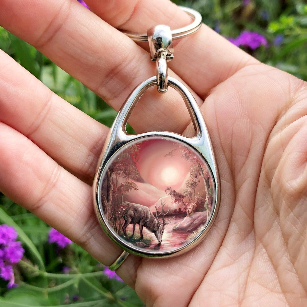 A stag standing on a river bank with his head down, taking a drink. The design is cast in shades of pink. Pink hills rise each side in the distance with the sun in the centre. A bird of prey hovers above. The banks of the stream are shallow in the foreground, but rise more steeply in the distance. Trees grow from the banks, with grass, flowers and toadstools on the shallower ones.