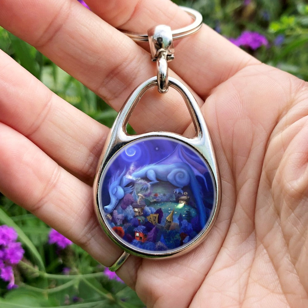 Painting of a blue dragon with rounded body shape and features sleeps on a hill, his head and tail draped down either side. Sheep graze on the hill. At the bottom of the hill lies a village with colourful trees, houses and castle. A crescent moon shines overhead.