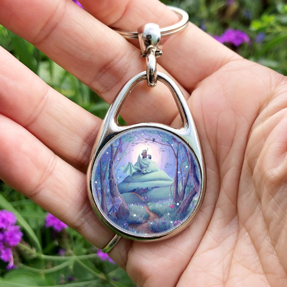 A winding path leads out from an enchanted forest where many fairies, and a dragonfly, can be seen. One fairy follows the path which leads up the side of a tall hill with steeply sloping sides. There's a tower on the top of the hill, and just below the tower, with its tail wound around it, sits a friendly dragon, wings partly open, and head resting on the path. There's a glow behind the top of the hill, fading to a blue night sky. Colours are rich teal, blue, purple and brown.