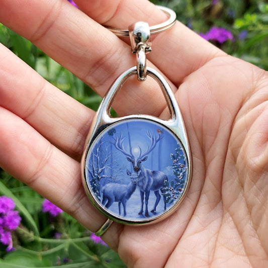 A stag and doe stand facing each other, heads turned towards the viewer. The design is cast in shades of blue. A robin perches on one of the stag's antlers. Snow lies underfoot, and on the branches of the berried holly tree to the right, and bare tree to the left. Pale fairies flit through the branches. A wintery moon rises from between the stag's antlers, and stars can be seen in the sky beyond.