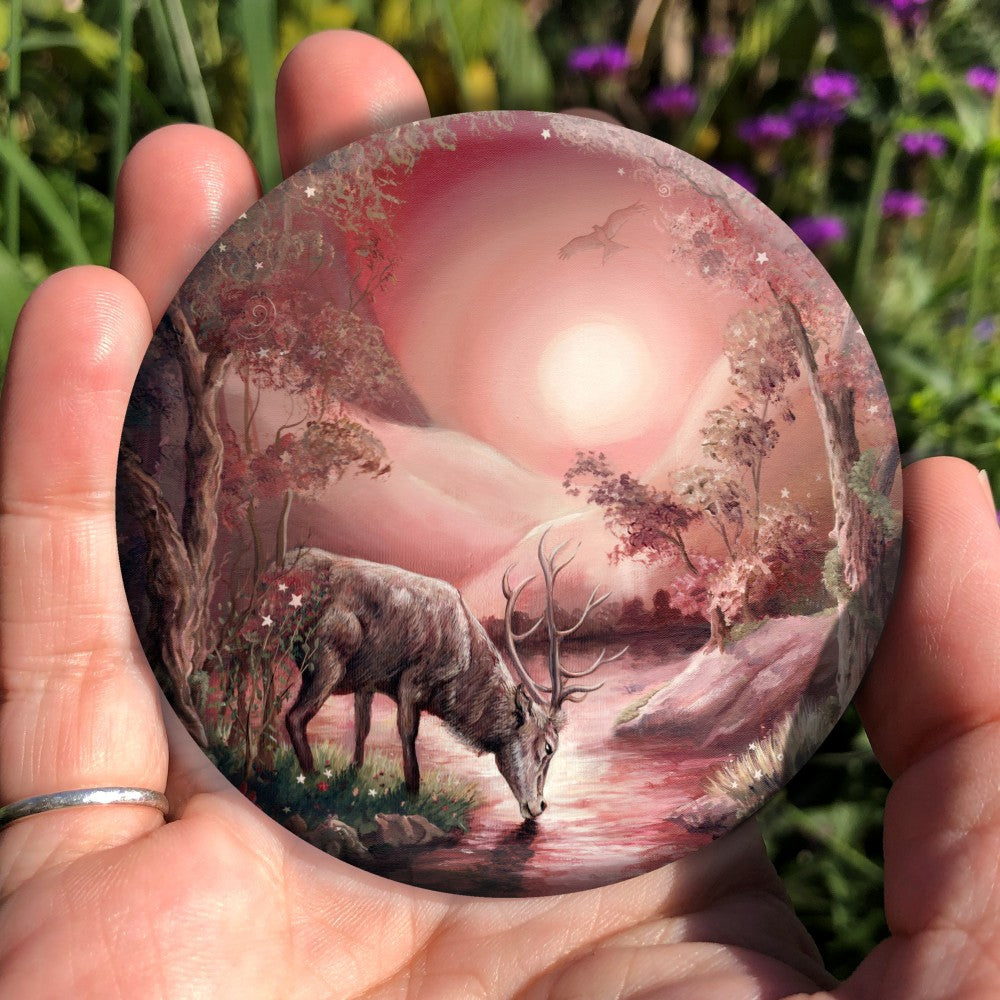 A stag standing on a river bank with his head down, taking a drink. The design is cast in shades of pink. Pink hills rise each side in the distance with the sun in the centre. A bird of prey hovers above. The banks of the stream are shallow in the foreground, but rise more steeply in the distance. Trees grow from the banks, with grass, flowers and toadstools on the shallower ones.