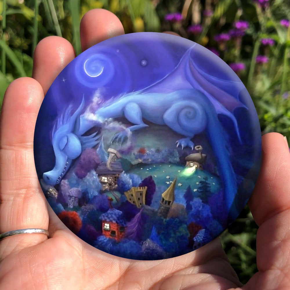 Painting of a blue dragon with rounded body shape and features sleeps on a hill, his head and tail draped down either side. Sheep graze on the hill. At the bottom of the hill lies a village with colourful trees, houses and castle. A crescent moon shines overhead.