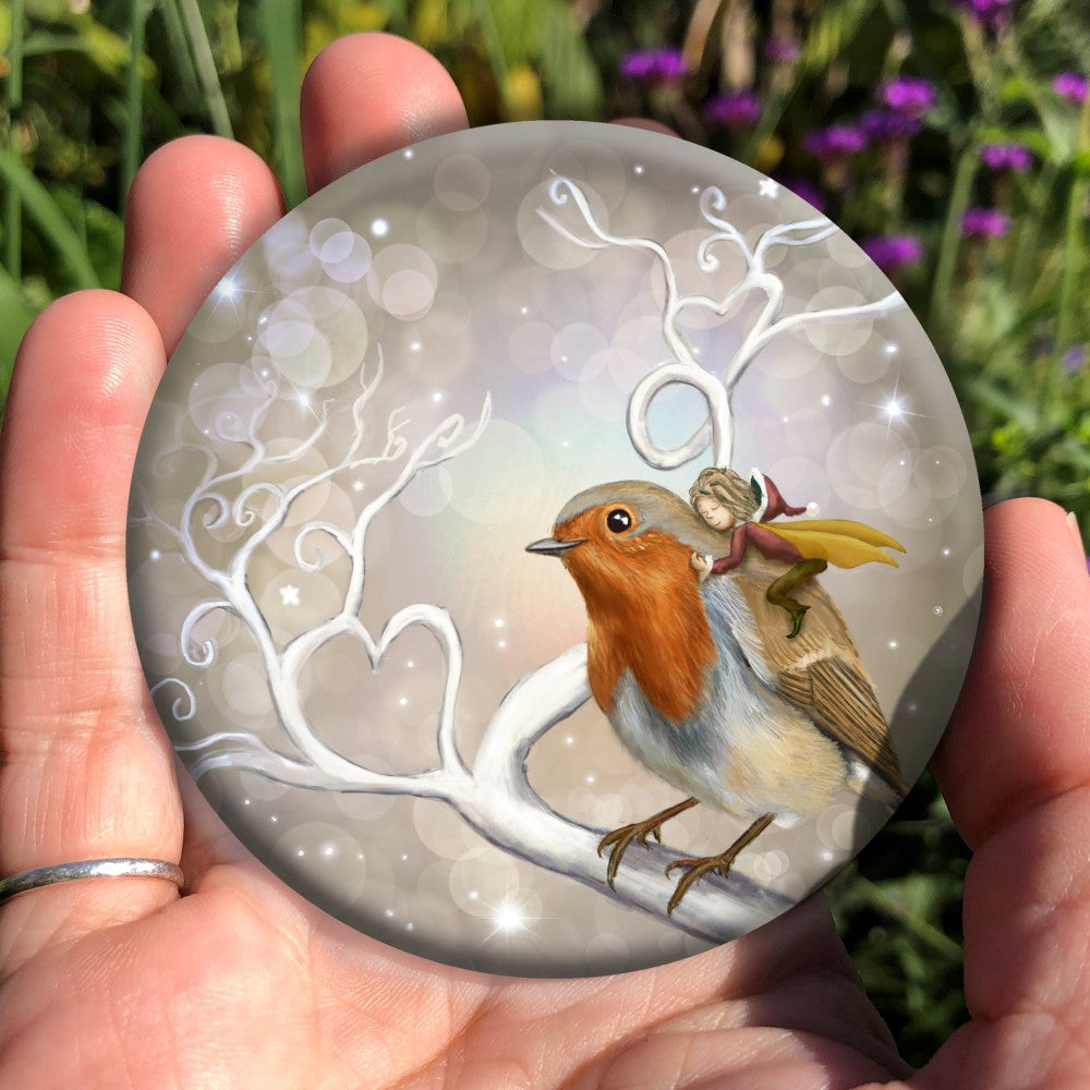 A robin perches on a white leafless branch. Twigs twist and curl from the branch, one forming a heart shape. A fairy with mustard-coloured wings and wearing red and brown, and a santa hat, holds onto the back of the robin, eyes closed and looking content. The background is mid-brown with stars and bubbles.