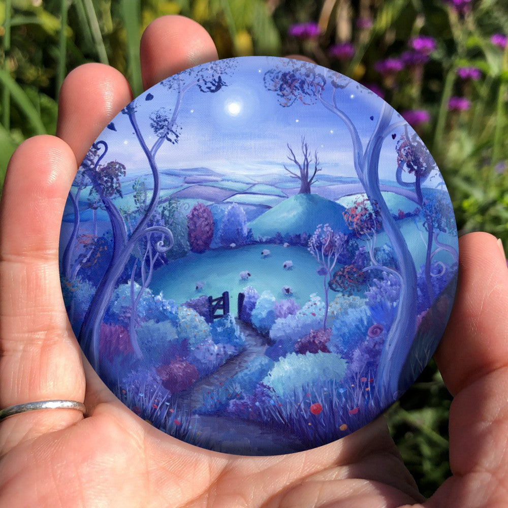 A path out of woodland leads away down a slope towards an open gate. Beyond the gate, sheep graze in a field, and beyond that there's a steep hill with a large dead tree on top. A patchwork of fields can be seen in the distance. A full moon shines in a mid-blue, starlit sky. The design is very colourful, with teal, purple, pink and blue hedges, trees, and fields.