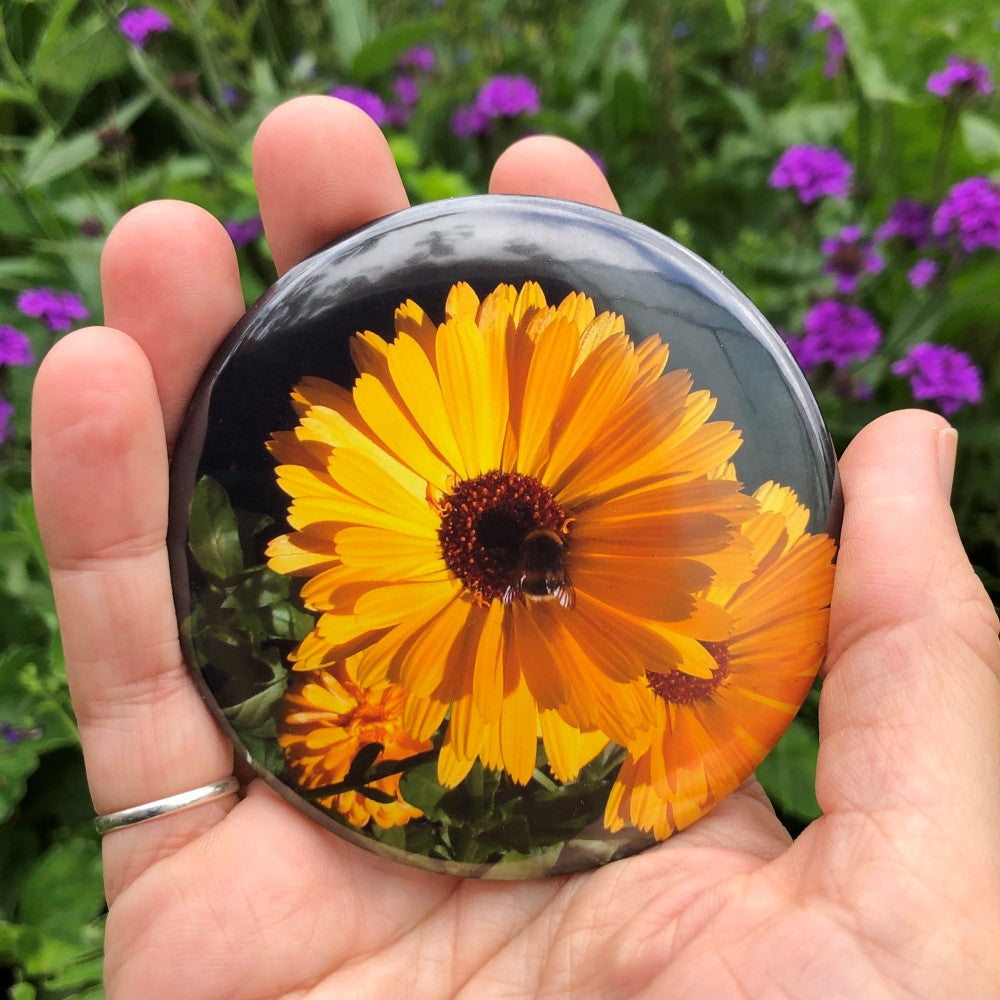 A bright orange flower with multiple layers of straight petals and a black centre is central to the design. A small bumble bee sits in the centre. More orange flowers and leaves can be seen in the background.