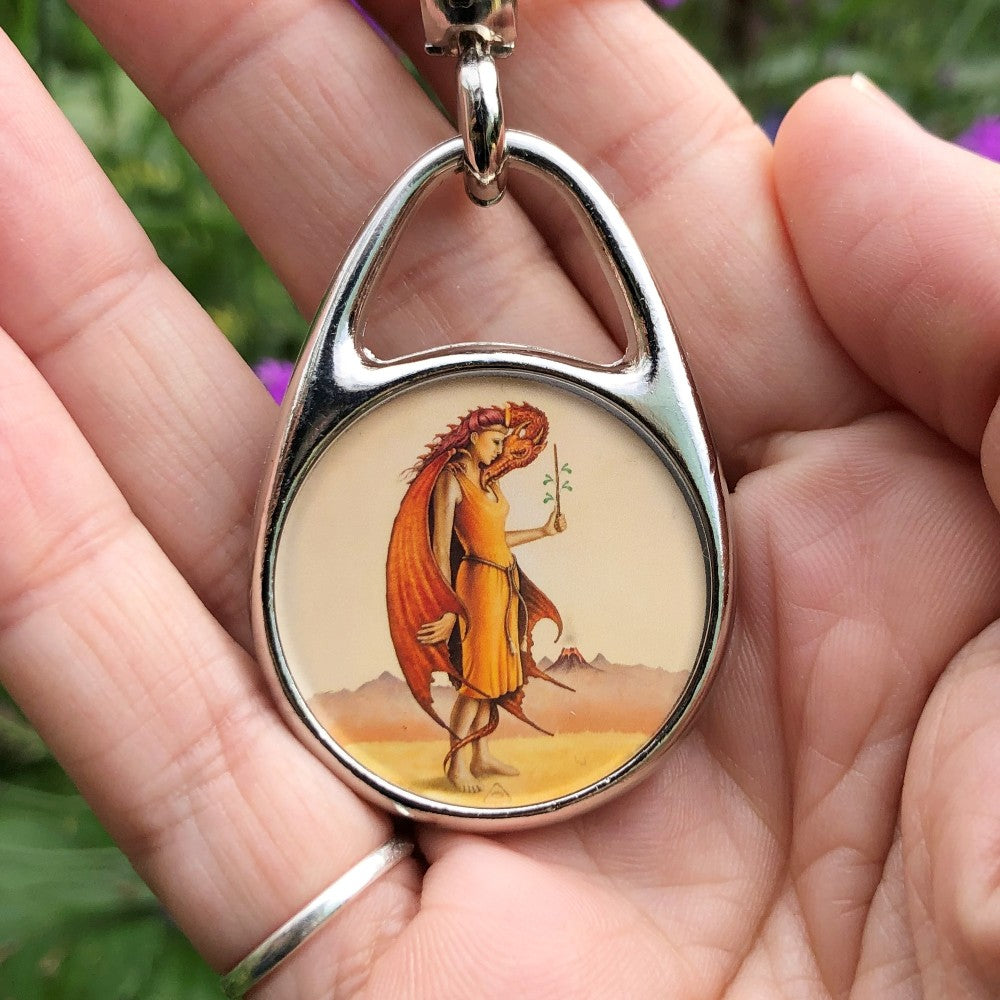 A woman in an orange dress and holding a wand upright in her left hand, faces to the right. Wrapped around her body with its head under her chin, is a deep orange dragon. She holds the edge of the dragon's wing in her right hand. In the distance, a volcano erupts. Colours are yellows and oranges.