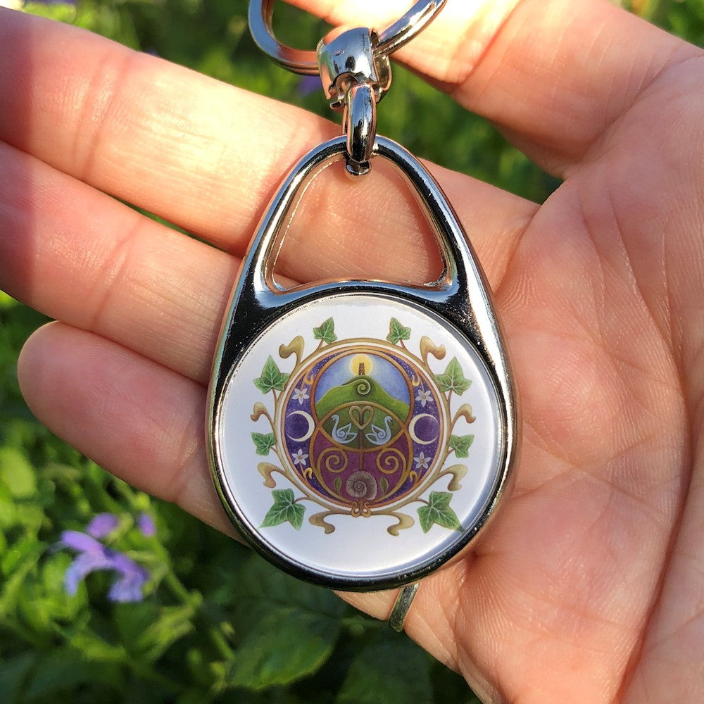 A circular design on a white background featuring the Chalice Well (Vesica Pisces) symbol inside a circle of ivy leaves. Glastonbury Tor, the sun rising behind the tower against a blue sky, nestles inside the upper circle of the symbol, with two swans in the area where the two circles join. An ammonite is inside the lower circle, against a maroon background. To the left and right are two opposing crescent moons, and small white flowers.