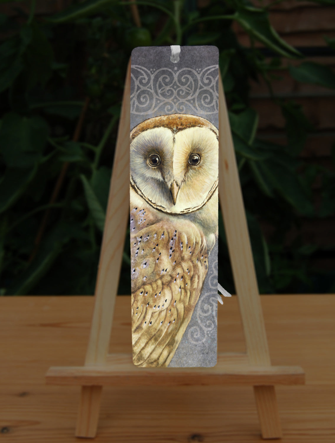 Rectangular bookmark: A pale brown bird with cream face sits sideways, coming in from the left hand side of the design, and looking directly at the viewer. The background is grey, overlaid a paler grey border consisting of a pattern of swirls.