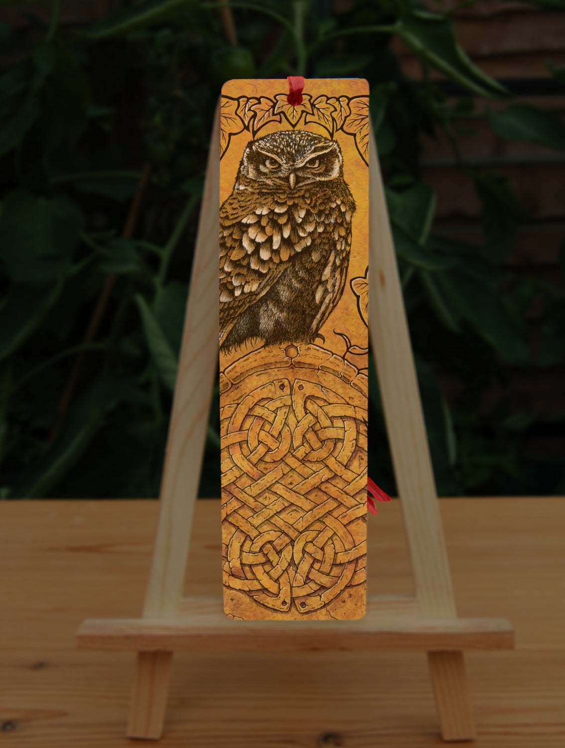Rectangular bookmark: A compact-shaped owl with brown feathers sits sideways on an upright, roughly semi-circular stone arch, while looking directly at the viewer. The arch is decorated with a central circular Celtic knotwork design. The design is mostly monochrome apart from the owl, with a background of mottled dark tan.