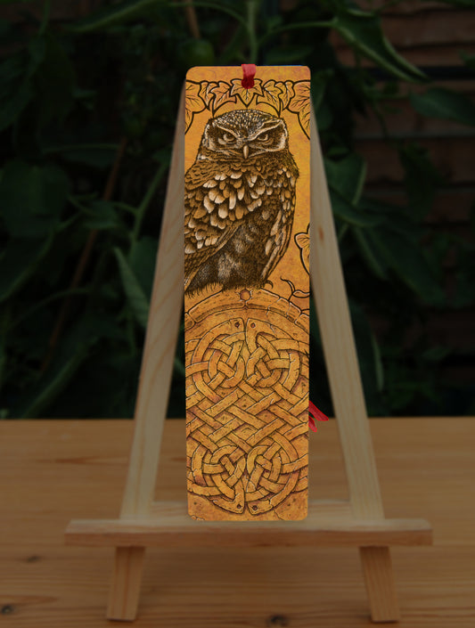 Rectangular bookmark: A compact-shaped owl with brown feathers sits sideways on an upright, roughly semi-circular stone arch, while looking directly at the viewer. The arch is decorated with a central circular Celtic knotwork design. The design is mostly monochrome apart from the owl, with a background of mottled dark tan.