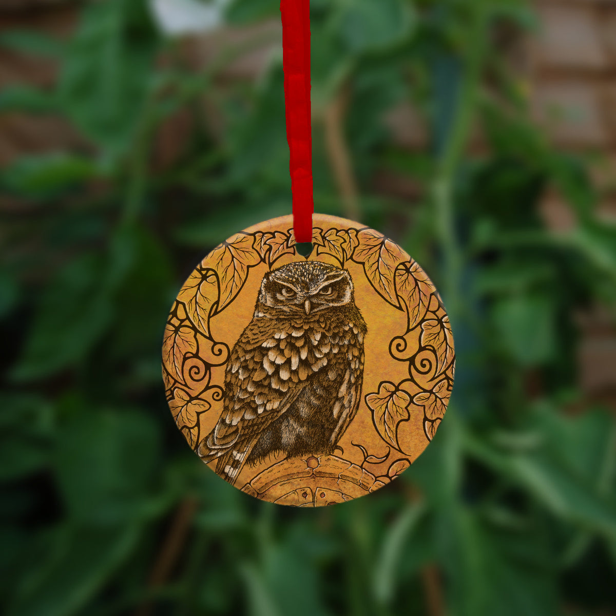 Circular hanging ornament with red ribbon: A compact-shaped owl with brown feathers sits sideways on an upright, roughly semi-circular stone arch, while looking directly at the viewer. A border of ivy leaves and simple curls surrounds the owl and down each side of the arch. The design is mostly monochrome apart from the owl, with a background of mottled dark tan.