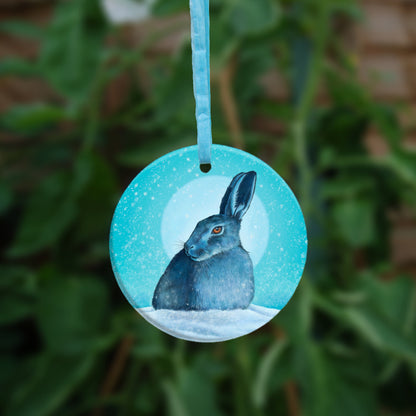 A hare crouches in snow, head turned to one side so one eye faces the viewer. Snow is falling, and a pale wintery sun shines behind. The design is coloured in shades of blue and turquoise, including the hare, which appears blue except for a deep orange eye.