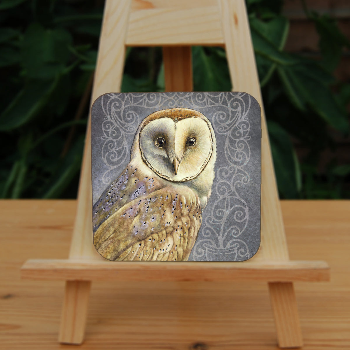 Square coaster: A pale brown bird with cream face sits sideways, coming in from the left hand side of the design, and looking directly at the viewer. The background is grey, overlaid a paler grey border consisting of a pattern of swirls.