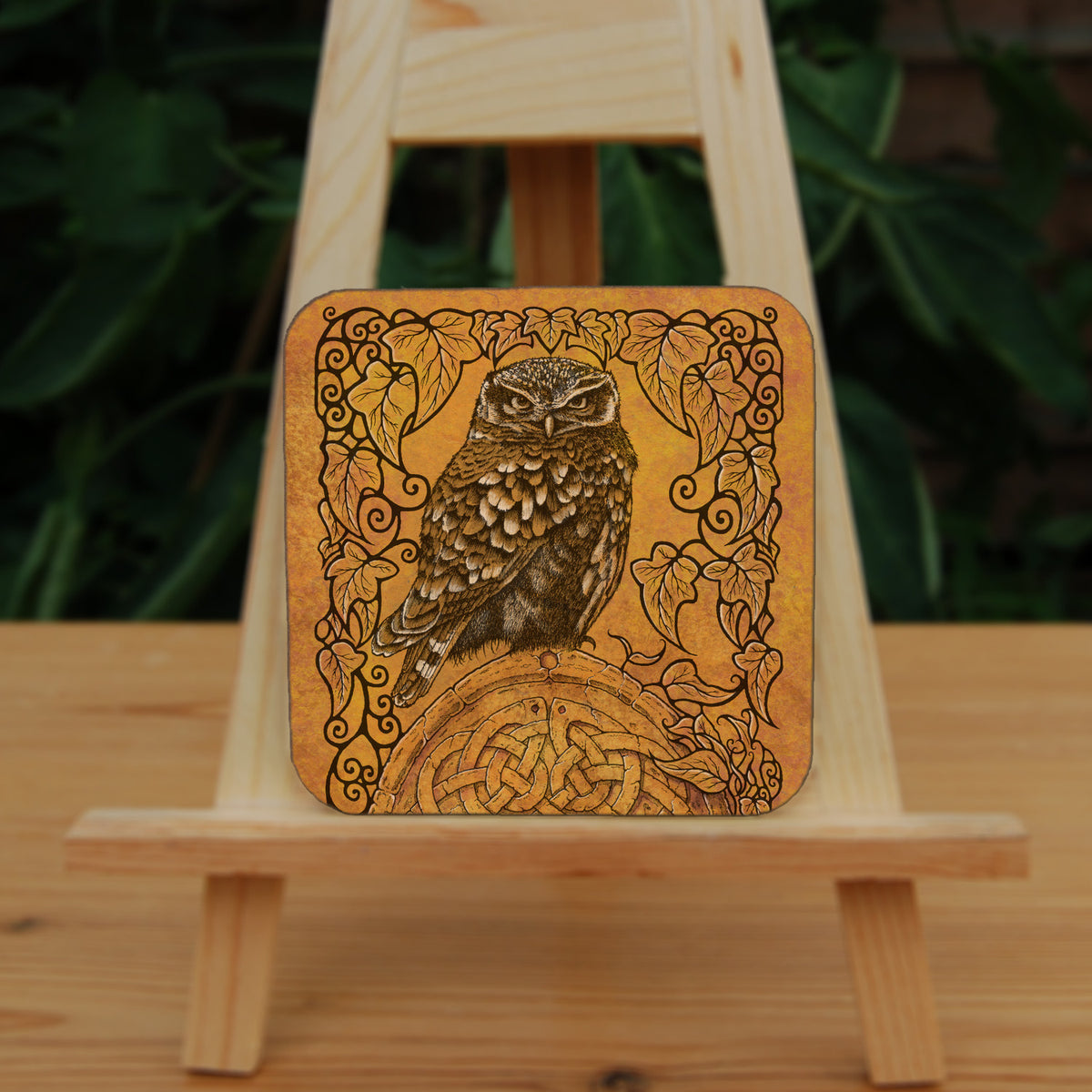 Square coaster: A compact-shaped owl with brown feathers sits sideways on an upright, roughly semi-circular stone arch, while looking directly at the viewer. The arch is decorated with a central circular Celtic knotwork design. A border of ivy leaves and simple curls surrounds the owl and down each side of the arch. The design is mostly monochrome apart from the owl, with a background of mottled dark tan.