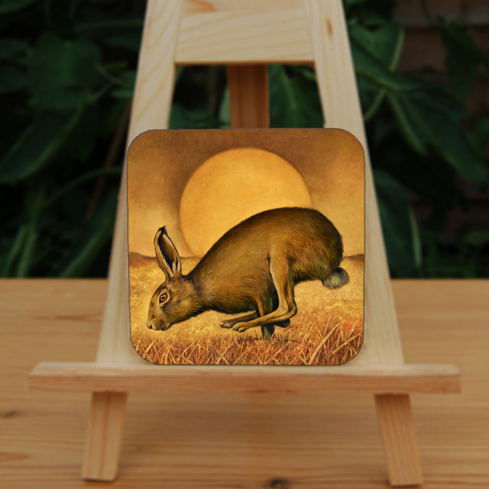 A brown hare runs from right to left, back curved, hind legs ahead of front legs. The sun rises behind, casting a golden brown glow across the sky, field and grass beneath the hare.