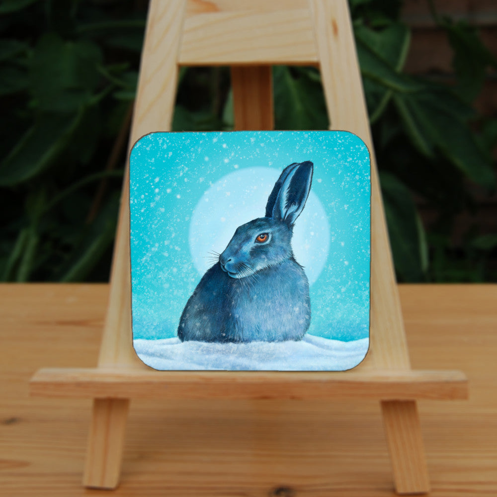 A hare crouches in snow, head turned to one side so one eye faces the viewer. Snow is falling, and a pale wintery sun shines behind. The design is coloured in shades of blue and turquoise, including the hare, which appears blue except for a deep orange eye.