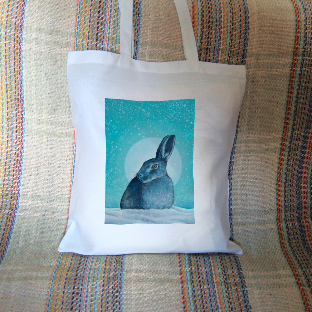 A hare crouches in snow, head turned to one side so one eye faces the viewer. Snow is falling, and a pale wintery sun shines behind. The design is coloured in shades of blue and turquoise, including the hare, which appears blue except for a deep orange eye.