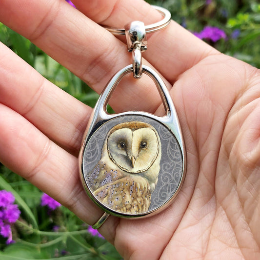 Hand holding a metal oval keyring with a circular insert: A pale brown bird with cream face sits sideways, coming in from the left hand side of the design, and looking directly at the viewer. The background is grey, overlaid a paler grey border consisting of a pattern of swirls.