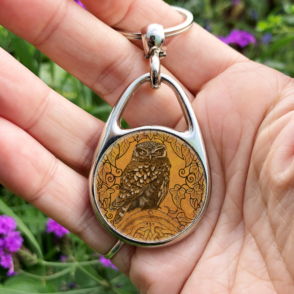 Hand holding a metal oval keyring with a circular insert: A compact-shaped owl with brown feathers sits sideways on an upright, roughly semi-circular stone arch, while looking directly at the viewer. The arch is decorated with a central circular Celtic knotwork design. A border of ivy leaves and simple curls surrounds the owl and down each side of the arch. The design is mostly monochrome apart from the owl, with a background of mottled dark tan.