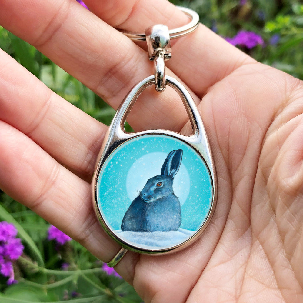 A hare crouches in snow, head turned to one side so one eye faces the viewer. Snow is falling, and a pale wintery sun shines behind. The design is coloured in shades of blue and turquoise, including the hare, which appears blue except for a deep orange eye.