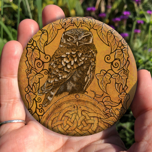 Hand holding a circular object: A compact-shaped owl with brown feathers sits sideways on an upright, roughly semi-circular stone arch, while looking directly at the viewer. The arch is decorated with a central circular Celtic knotwork design. A border of ivy leaves and simple curls surrounds the owl and down each side of the arch. The design is mostly monochrome apart from the owl, with a background of mottled dark tan.