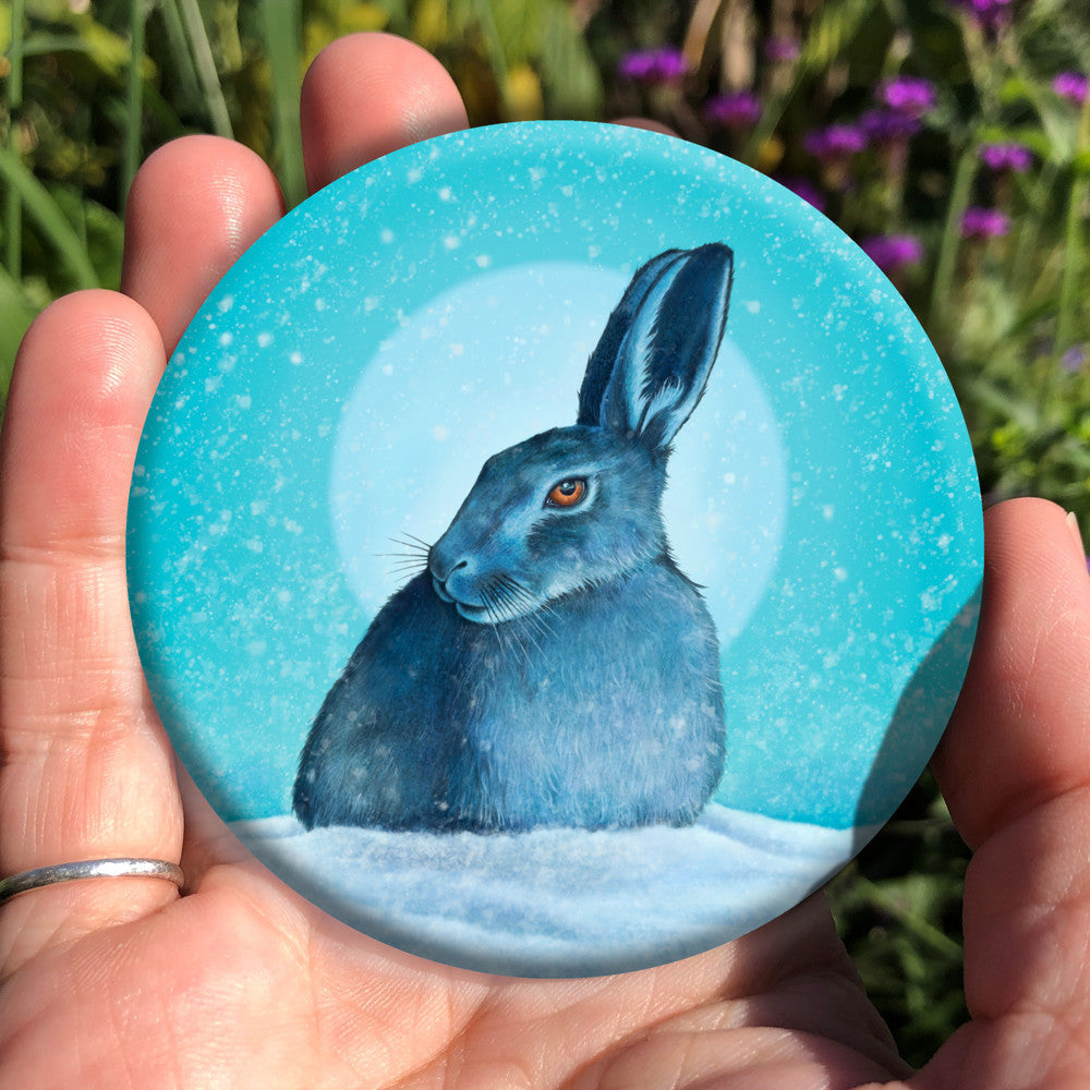 A hare crouches in snow, head turned to one side so one eye faces the viewer. Snow is falling, and a pale wintery sun shines behind. The design is coloured in shades of blue and turquoise, including the hare, which appears blue except for a deep orange eye.