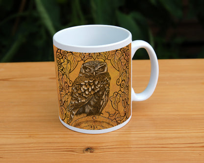 A white ceramic mug: A compact-shaped owl with brown feathers sits sideways on an upright, roughly semi-circular stone arch, while looking directly at the viewer. A border of ivy leaves and simple curls surrounds the owl and down each side of the arch. The design is mostly monochrome apart from the owl, with a background of mottled dark tan.