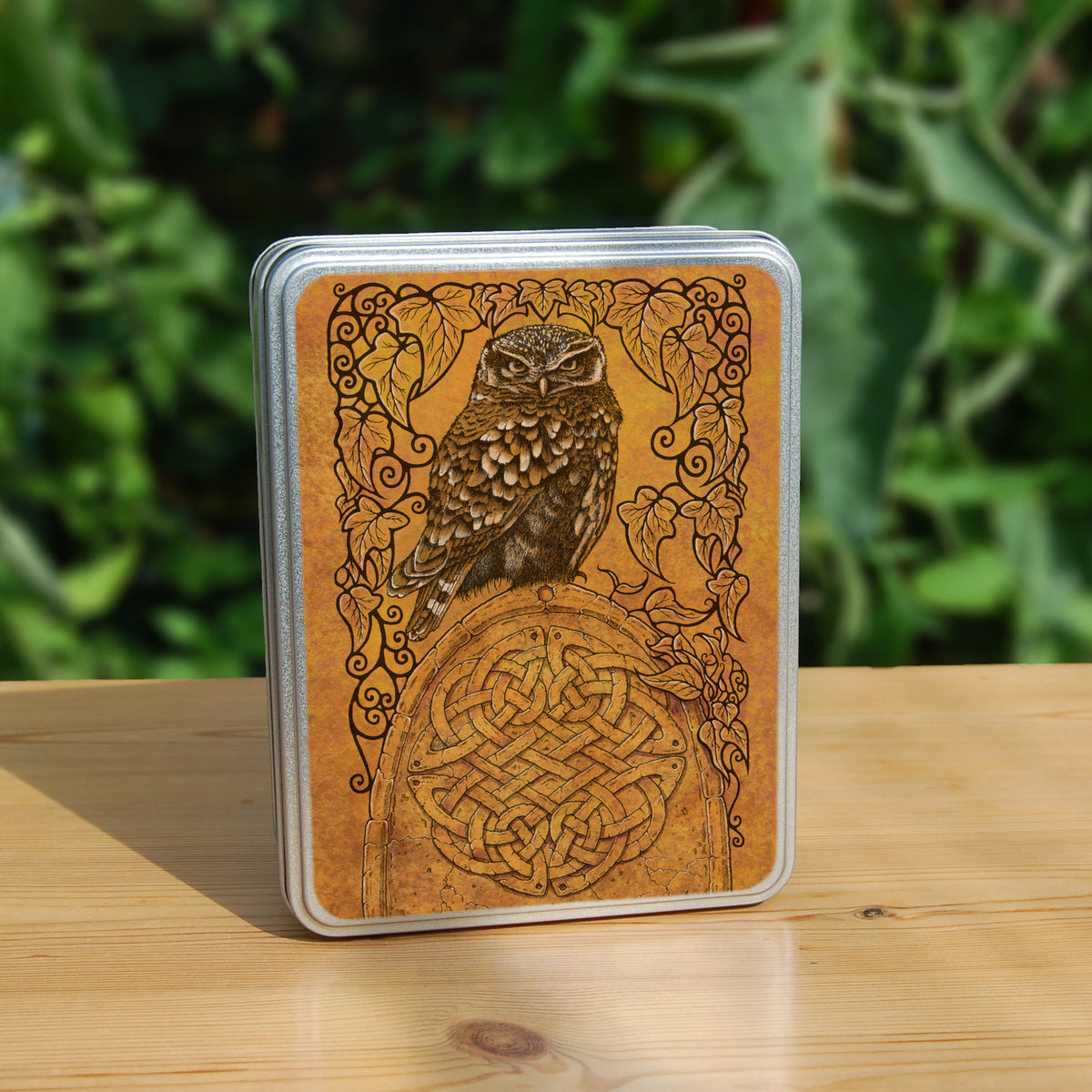 A rectangular metal tin: A compact-shaped owl with brown feathers sits sideways on an upright, roughly semi-circular stone arch, while looking directly at the viewer. The arch is decorated with a central circular Celtic knotwork design. A border of ivy leaves and simple curls surrounds the owl and down each side of the arch. The design is mostly monochrome apart from the owl, with a background of mottled dark tan.