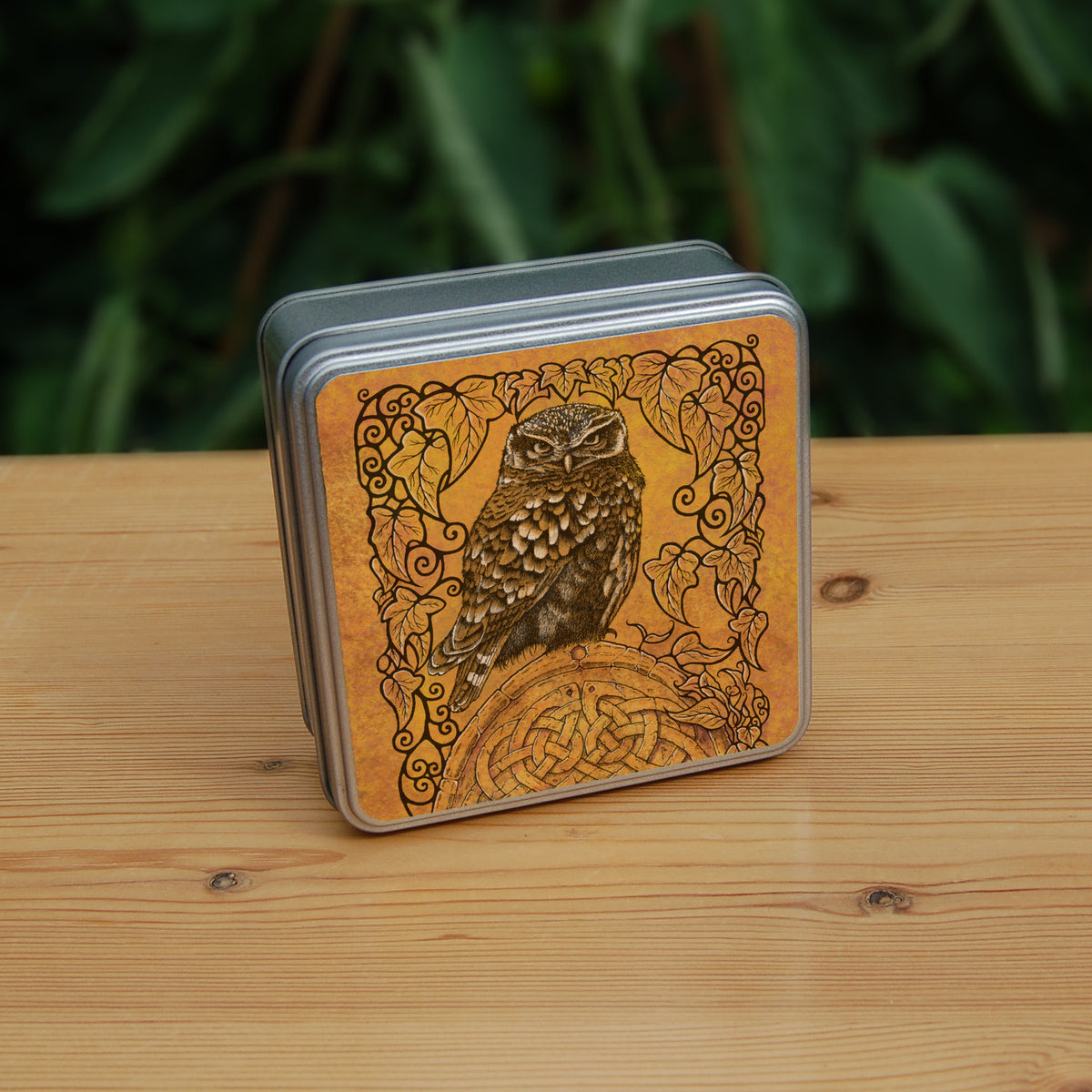 Square metal tin: A compact-shaped owl with brown feathers sits sideways on an upright, roughly semi-circular stone arch, while looking directly at the viewer. The arch is decorated with a central circular Celtic knotwork design. A border of ivy leaves and simple curls surrounds the owl and down each side of the arch. The design is mostly monochrome apart from the owl, with a background of mottled dark tan.